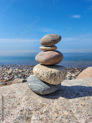 Tranquil Scene of a Stack of Smooth and Round Stones of Various Sizes Arranged on the Rocky Shore of a Calm Blue Lake, Set Against a Cloudless Sky Background in Bright Sunlight