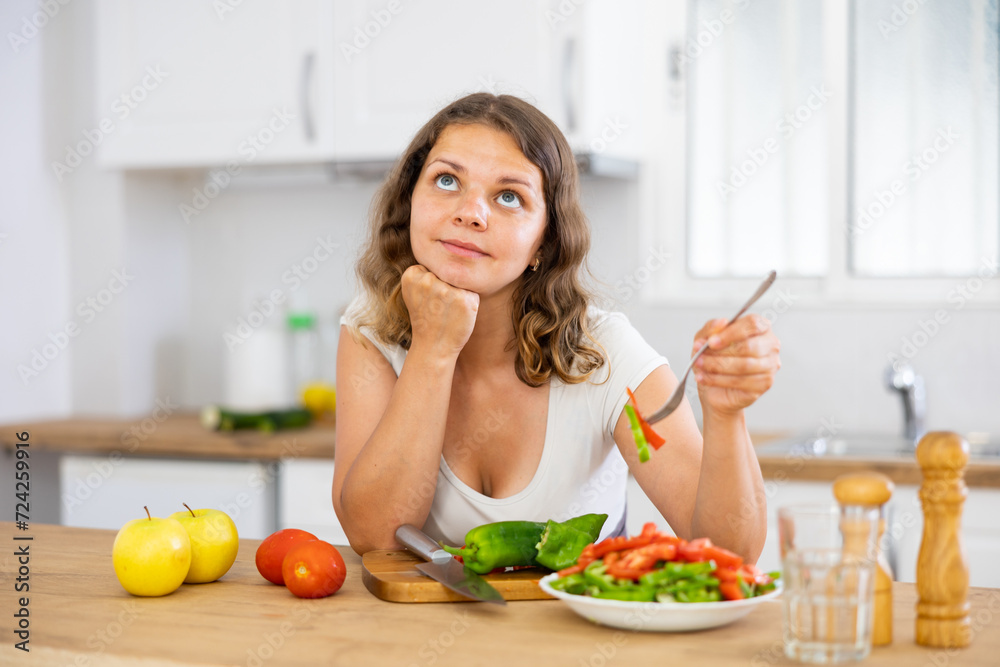 Portrait of thoughtful woman standing at kitchen table and eating just cooked fresh salad.