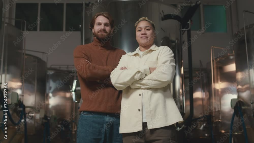 Low angle medium portrait of two confident ethnically diverse male and female beer technicians posing for camera with hands folded at brewing factory Low angle medium portrait of two confident ethnically diverse male and female beer technicians posing for camera with hands folded at brewing factory