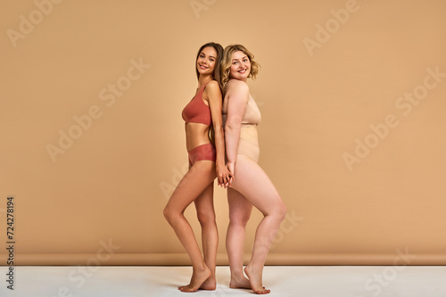 Diverse beauty. Two caucasian women with various body size and skin color standing close and smiling over beige background. Female friends wearing comfortable lingerie holding hands in support.