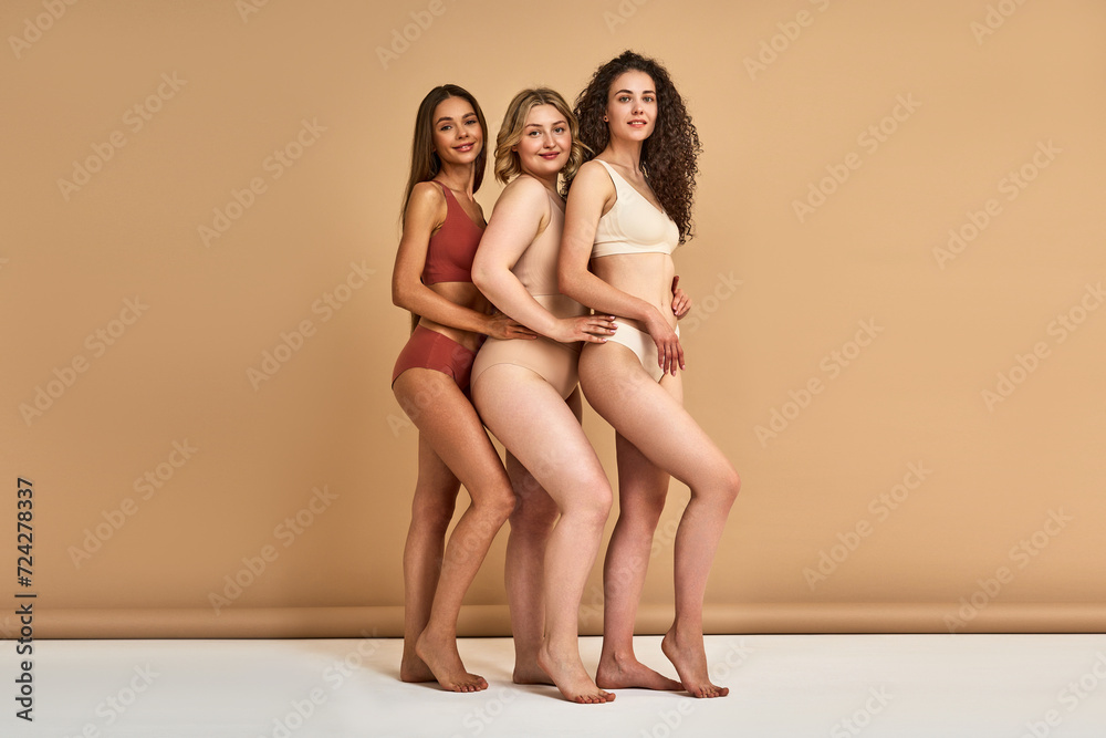 Diverse female beauty. Group of three attractive women with different skin color and body shape posing in comfy underwear over beige background. Self-acceptance and self-love concept.
