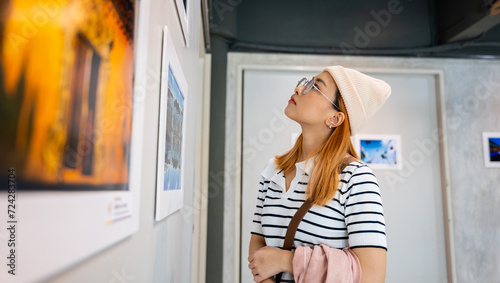 Woman standing she looking art gallery collection in front framed paintings pictures on white wall, Asian people watch at photo frame to leaning against at show exhibition artwork gallery, Side view photo
