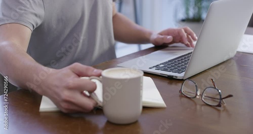 Man, laptop and hands typing while drinking coffee for morning in remote work on desk at home. Closeup of male person or freelancer working on computer with cup of tea and book for email at house photo