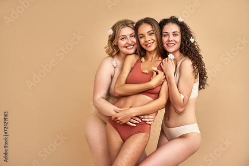 International Women's Day. Pleasant women with various size smiling at camera while posing in studio with flowers on tender bodies. Charming female wearing comfy red, beige and white sports underwear photo