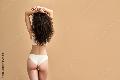 Expression of self love. Back view of slender caucasian woman with arms raised up posing sensually over beige background. Charming young lady having long curly hair and wearing comfy soft underwear. photo