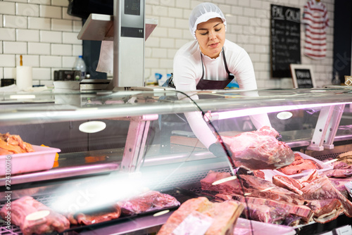 Positive young butcher shop saleswoman arranging meat products on display case, showing piece of fresh raw veal tenderloin