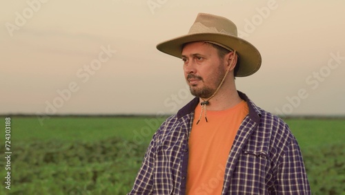 Long man farmer smiles. Portrait of farmer looking at camera, adjusting his hat. Slow motion. Closeup of handsome young business man with beard smiling at camera, green field. Agricultural business