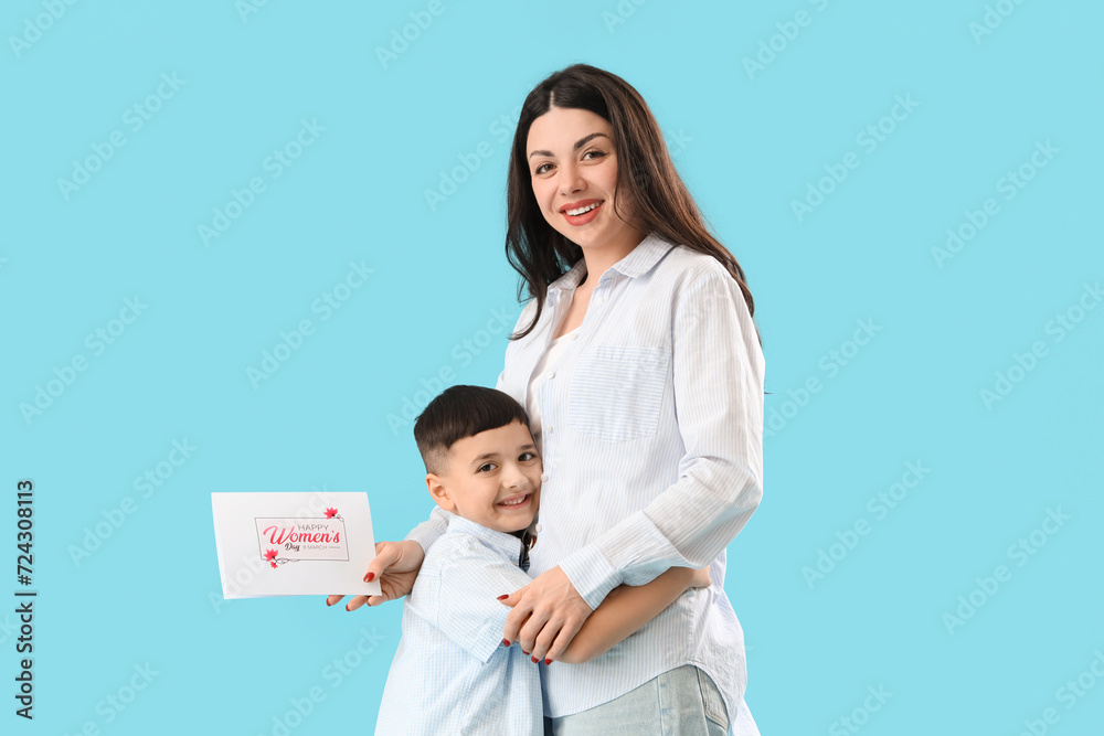 Beautiful woman with greeting card and her little son on blue background. International Women's Day