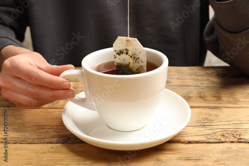 Woman putting tea bag in cup with hot water at wooden table, closeup