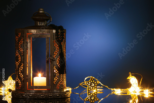 Muslim lamp with burning candle, garland and prayer beads for Ramadan on dark blue background