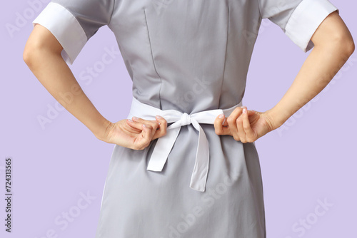 Young chambermaid tying apron on lilac background, back view