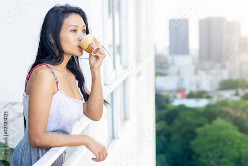A woman in a summer dress drinks from a teacup on the balcony of an apartment building above city park