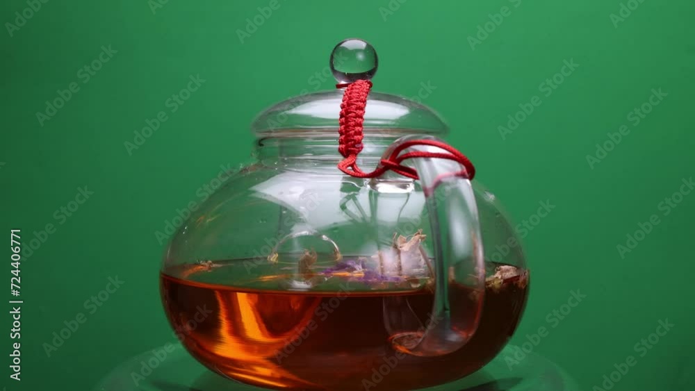 Rotating transparent glass teapot brewing fresh herbal flower tea in hot water on green background