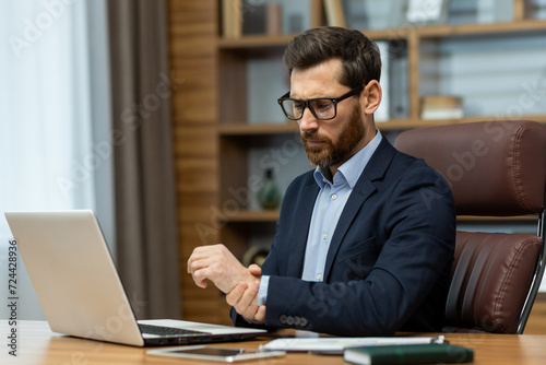 Tired entrepreneur in formal clothes massaging wrist with hand after long day of using laptop. Successful copywriter having spasms in palm muscles after typing on pc and answering emails in workplace.