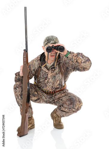 Portrait of squatting mature hunter with a shotgun looks through binoculars to the camera, isolated on white background. Duck hunter with a rifle posing in studio.
