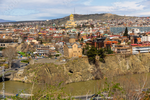 Scenic cityscape of historical area of Tbilisi overlooking Holy Trinity Cathedral with golden dome in background and small Metekhi Church on steep bank of Mtkvari River on sunny spring day, Georgia