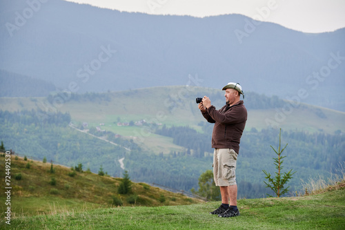 Male tourist hiking in hills, traveling in summer. Man standing on hill, holding camera, taking photo, admiring landscape, enjoying. Concept of tourism and adventure.