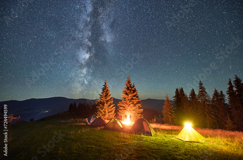 Night camping in mountains under starry sky. Tents standing on grass in campsite, fire burning bright, stars shining on sky. Concept of tourism and adventure.