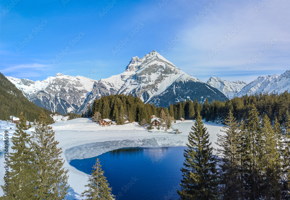 Aerial image of the winter landscape of the frozen Arni Lake with the snow-convered Alps peak Windgaellen at the background - obrazy, fototapety, plakaty 