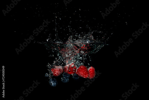 A handful of raspberries and blueberries falling into water with a black background creating bubbles and a splash