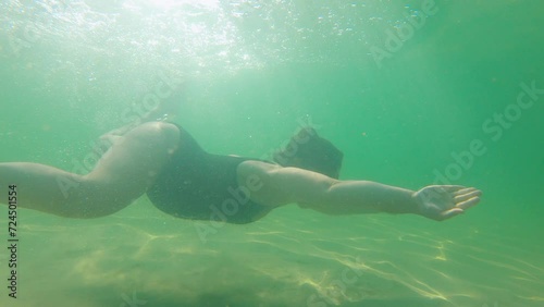 Pregnant, beautiful woman in a black swimsuit swims underwater in the tropical sea, in which the rays of the sun play. Healthy and active pregnancy concept. Water activities, travel during pregnancy
