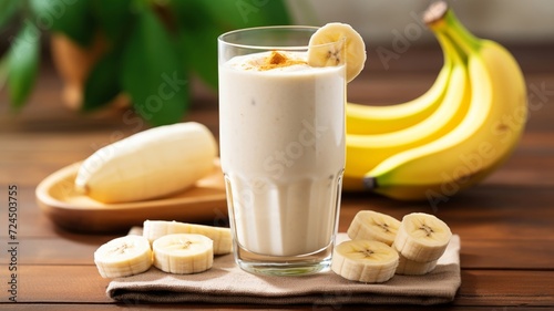 Creamy banana milkshake topped with a slice of banana, presented on a white distressed wood table with fresh bananas in the background