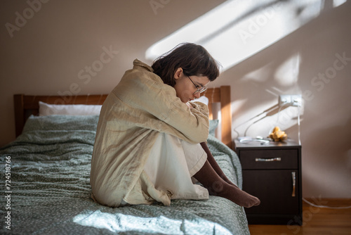Indifferent unemotional unsociable teenager sitting on bed embracing knees alone at home, looking desperately on floor. Friendlessness, solitude, loss of life interest, self destruction concept photo