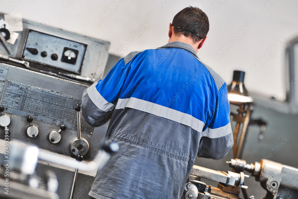 Turner in overalls works at lathe in turning shop at factory. Elderly worker turns metal part on machine. Rear view from back. Turner in overalls works at lathe in turning shop at factory. Elderly worker turns metal part on machine. Rear view from back.