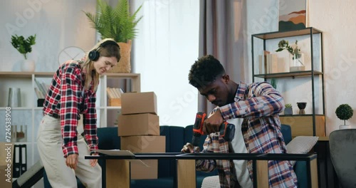 Attractive man twisting screw using handheld screwdriver assembling rack when his pretty positive relaxed woman listening music in headphones stacking cartons