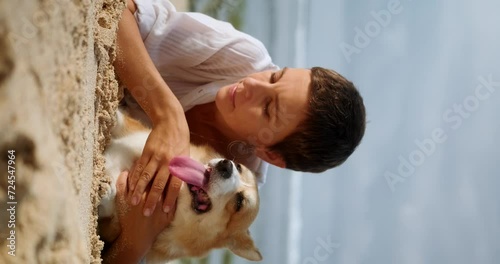 A middle-aged woman in a white shirt with short hair hugs and plays with her Welsh Corgi on the beach. The dog lies on the sand with a funny muzzle and his tongue hanging out. Traveling with a pet. photo