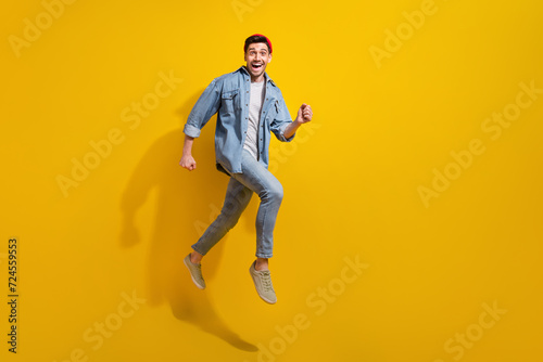 Full length photo of carefree impressed man dressed jeans shirt jumping high running fast empty space isolated yellow color background