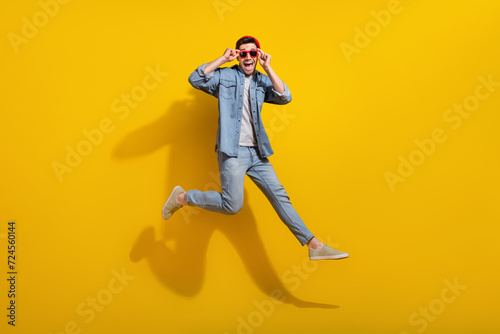 Full length photo of positive excited guy wear denim jacket dark glasses jumping high isolated yellow color background