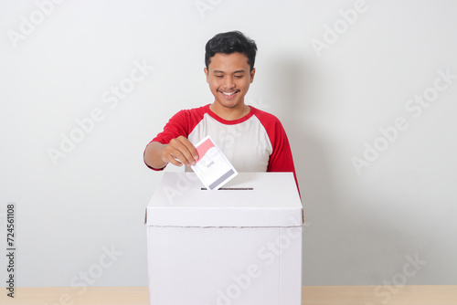 Portrait of excited Asian man inserting and putting the voting paper into the ballot box. General elections or Pemilu for the president and government of Indonesia. Isolated image on white background photo