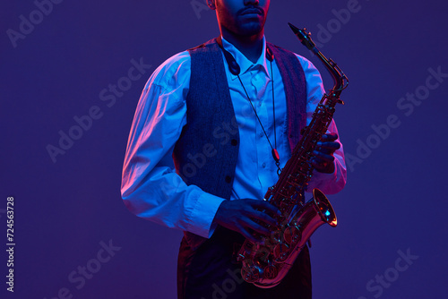 Canvas Print Portrait of young man, jazz performer, instrumental musician holds saxophone and looking at camera against dark background in neon light