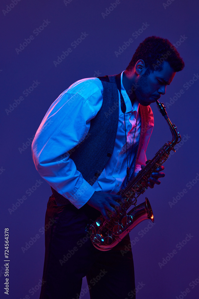 Naklejka premium African-American man in shirt and vest playing saxophone against blue and purple background in neon lighting. Concept of jazz, blues, classy instrumental music, festivals and concerts. Ad