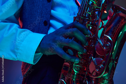 African-American artist, solo performer plays chords on fingering of saxophone, wind instrument in neon light against studio background. Concept of classical musical instrument, concert, festival. Ad photo