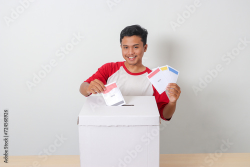 Portrait of excited Asian man inserting and putting the voting paper into the ballot box. General elections or Pemilu for the president and government of Indonesia. Isolated image on white background photo