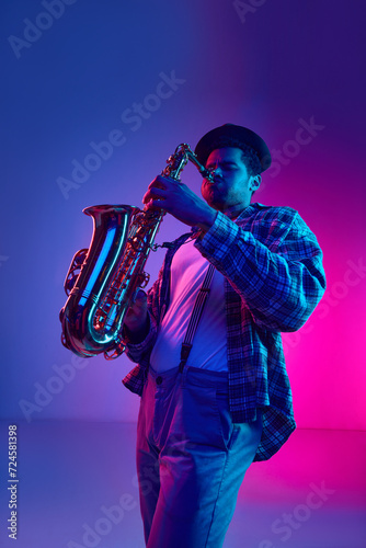 Jazz musician in retro outfit virtuously playing saxophone against gradient blue-pink background in neon light. Concept of blues, classy instrumental music, festivals and concerts. Ad