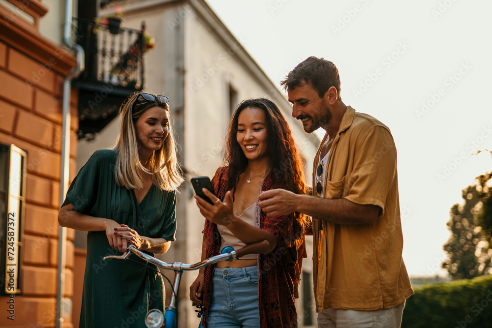 Three friends of different ethnicities, casually leaning on a bicycle, immersed in their smartphone while exploring the city