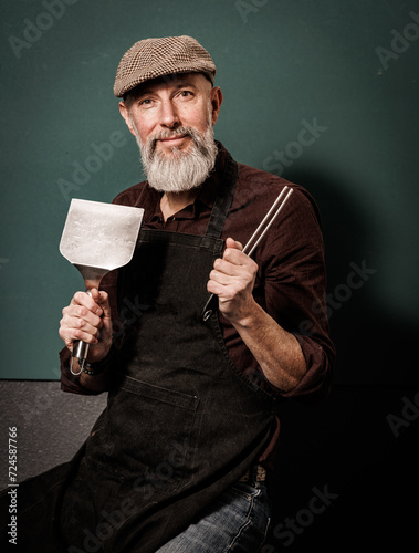 Portrait d'un homme barbu senior agé quinquagénaire hipster cuisinier qui tient des ustensiles de cuisine pour faire une plancha ou un barbenue photo