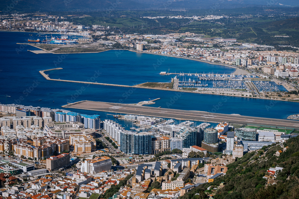 Gibraltar, Britain - January 24, 2024 - An aerial view of a densely built coastal city with a marina, an airport runway near the sea, industrial areas, and mountains in the background.