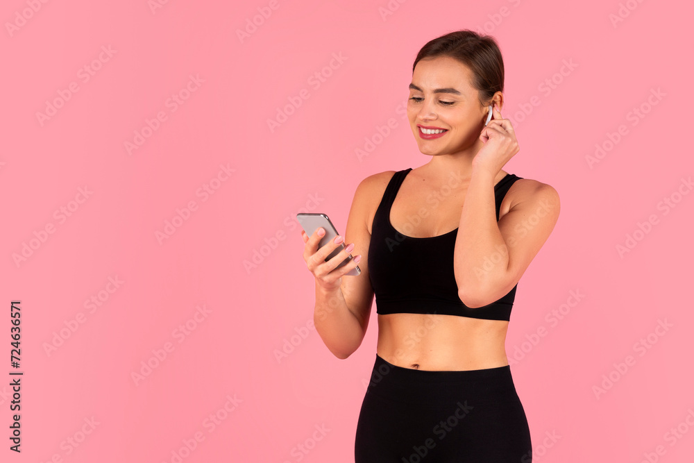Smiling young sportswoman adjusting wireless earbuds while looking at her smartphone Smiling young sportswoman adjusting wireless earbuds while looking at her smartphone
