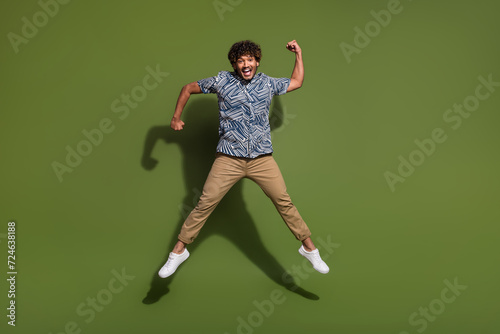 Full length photo of lucky impressed man dressed print shirt jumping high rising fist isolated green color background