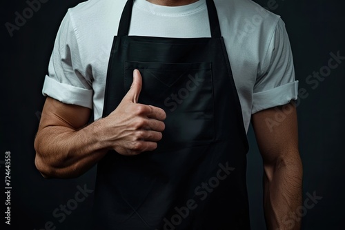 A powerful man in a black apron points towards the apron s center Implies chef or waiter Background is dark