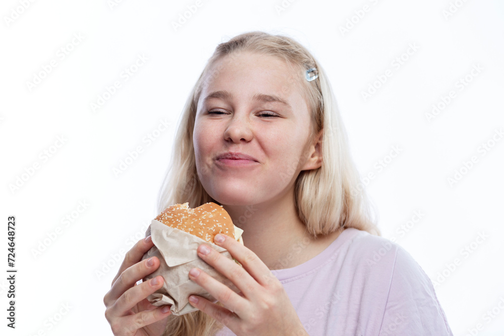 Smiling hungry teenage girl takes a bite of hamburger. Delicious popular fast food. A cute blonde with freckles in a pink jacket. White background.