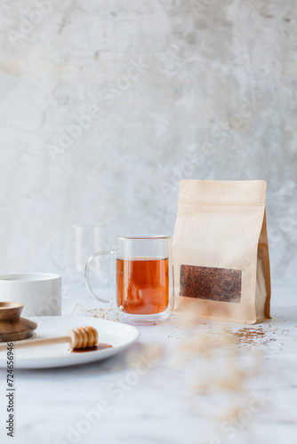 A styled lifestyle shot of a glass mug with black brewed Rooibos tea, surrounded by elements such as brown packaging with dried loose leaf tea, honey and a plate