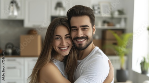 A young, happy couple, smiling, standing in white modern city apartment interior, surrounded by moving boxes on background