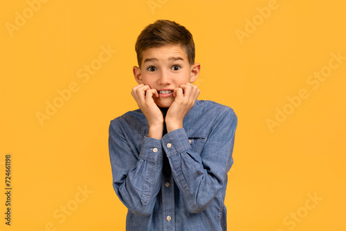 Anxious teenage boy in denim shirt biting nails and looking at camera