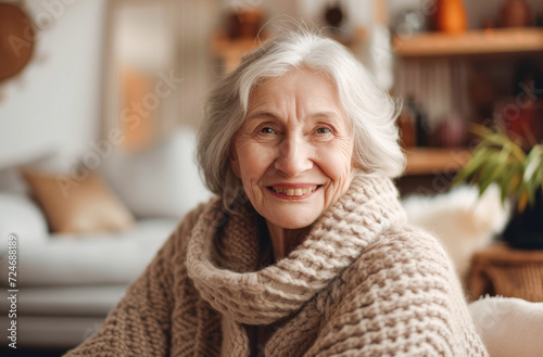 Portrait of beautiful mature woman smiling while sitting at sofa at home. Smiling retired grey haired woman looking at camera in winter time. Positive senior female sitting on sofa in living room. 