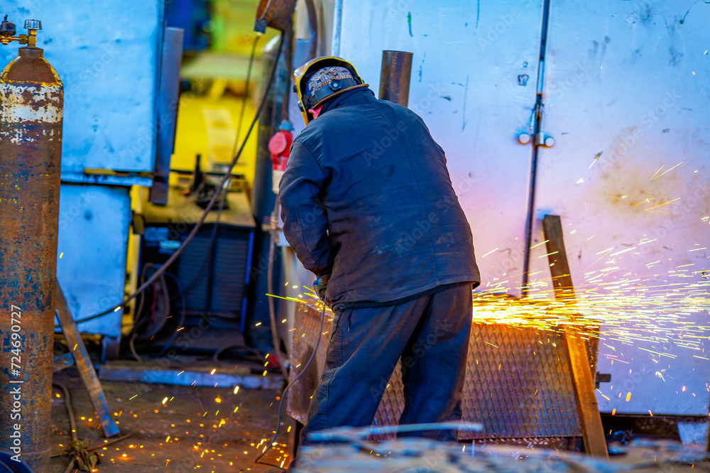 A worker cuts metal structures and parts with an abrasive tool at a factory. The grinder sparks when cutting steel. - obrazy, fototapety, plakaty 